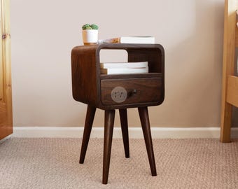 May include: A dark brown wooden nightstand with a drawer and three angled legs. The nightstand has a rounded top and a shelf. A small potted plant and books are on top. The nightstand is approximately 24 inches tall.