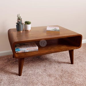 May include: A mid-century modern style wooden coffee table with rounded edges and tapered legs. The table has a lower shelf and is holding a book, a small succulent plant, and a small cactus plant.