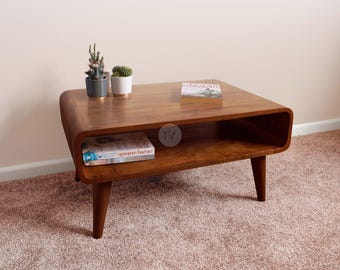 May include: A mid-century modern style wooden coffee table with rounded edges and tapered legs. The table has a lower shelf and is holding a book, a small succulent plant, and a small cactus plant.