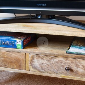 May include: A wooden TV stand with two drawers and a shelf. The stand has a light brown finish and is decorated with a distressed look. The top shelf has a board game box with the words "MONOPOLY" and "HOBBIT" on it. The bottom shelf has a book with the words "THE ARCHE OF STEPHENOISE" on it.