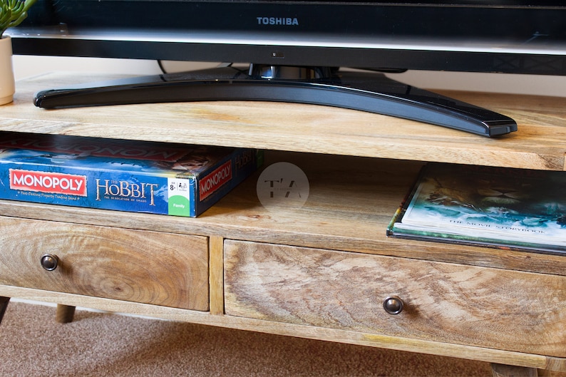 May include: A wooden TV stand with two drawers and a shelf. The stand has a light brown finish and is decorated with a distressed look. The top shelf has a board game box with the words "MONOPOLY" and "HOBBIT" on it. The bottom shelf has a book with the words "THE ARCHE OF STEPHENOISE" on it.