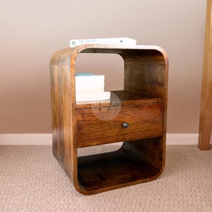 May include: A wooden bedside table with a curved design and a single drawer. The table has a shelf for books or other items. The table is made of dark wood and has a natural finish.