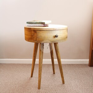 May include: A round wooden side table with a single drawer and tapered legs. The table is made of light brown wood and has a natural finish. The drawer has a black knob.