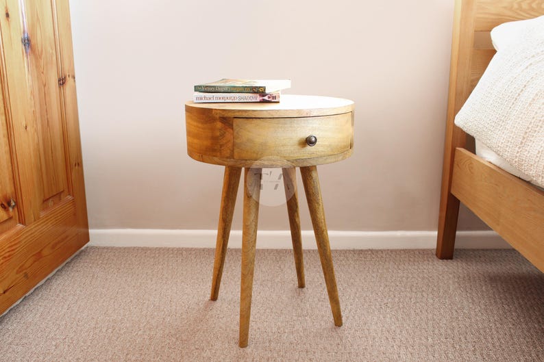 May include: A round wooden side table with a single drawer and tapered legs. The table is made of light brown wood and has a natural finish. The drawer has a black knob.