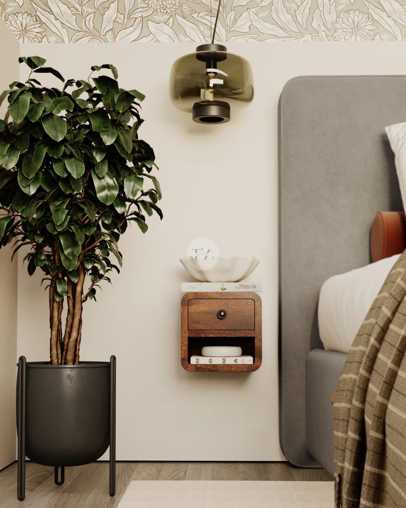 May include: A bedroom scene featuring a large potted plant with dark green leaves, a hanging glass lamp, and a floating wooden nightstand with a drawer. The nightstand has a small bowl and books on top. The bed has a gray headboard.