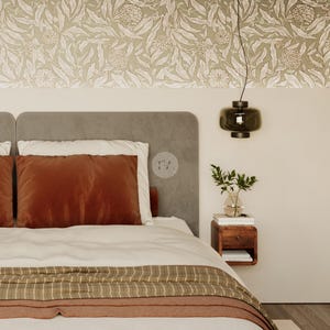 May include: A bedroom scene featuring a bed with a rust-colored velvet pillow and a striped throw blanket. The headboard is gray, and the wall has a floral wallpaper design. A dark green pendant light hangs above a wooden nightstand with a vase of greenery.