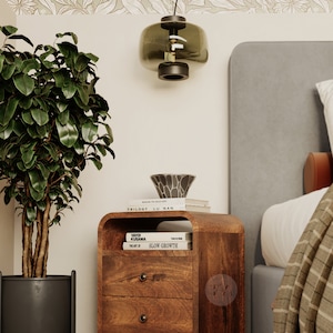 Pu&ograve; includere: Un comodino in legno marrone scuro con due cassetti e uno scaffale. Il comodino ha un bordo anteriore curvo ed &egrave; decorato con pomelli in ottone. Il comodino &egrave; accanto a un letto con una testiera grigia e una coperta bianca. Ci sono tre libri sopra il comodino, tra cui "Slow Growth" e "Yayoi Kusama".
