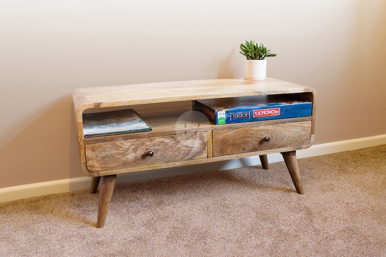 May include: A wooden TV stand with two drawers and a shelf. The stand has a curved front edge and is supported by four legs. The stand is made of light wood and has a natural finish. The stand is decorated with a plant in a white pot and a board game box on the shelf.