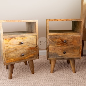 May include: Two wooden nightstands with two drawers each. The nightstands have a shelf above the drawers and are on short legs. The nightstands are made of light brown wood with a natural finish.