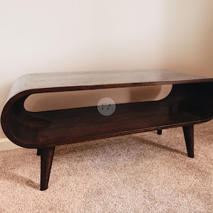 May include: A dark brown, mid-century modern style coffee table. The table has a rounded, oval shape with a lower shelf and four tapered legs. The table is made of wood and sits on a light brown carpet.
