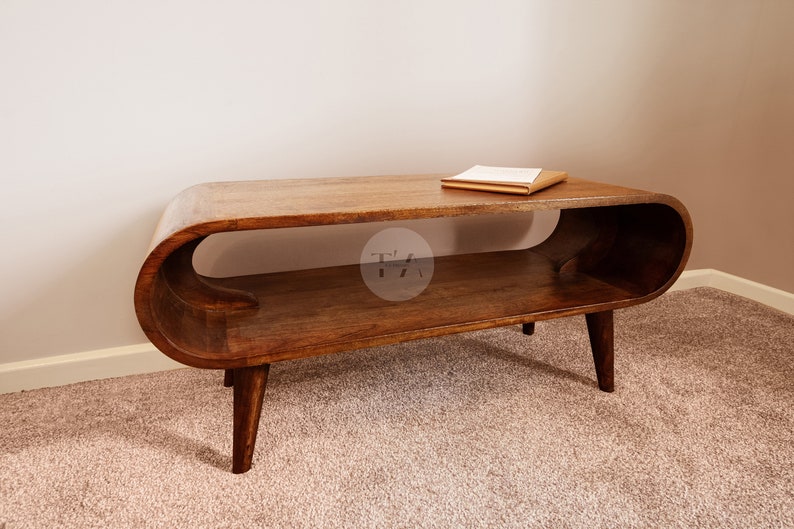 Op de afbeelding: Een houten salontafel met een uniek gebogen ontwerp en een lager plank. De tafel heeft een lichtbruine afwerking en staat op een beige tapijt.