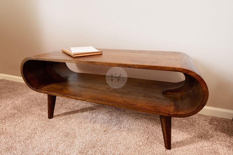 Op de afbeelding: Een donkerbruine houten salontafel met een gebogen, ovaalvormig blad en een lager plank. De tafel heeft vier poten, waarvan er twee zichtbaar zijn op de afbeelding. Een boek ligt op de bovenkant van de tafel.