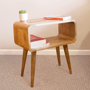 Peut inclure: Une table d'appoint en bois avec un design incurvé et un plateau blanc. La table a une étagère et quatre pieds. Il y a des livres et une plante en pot sur la table.