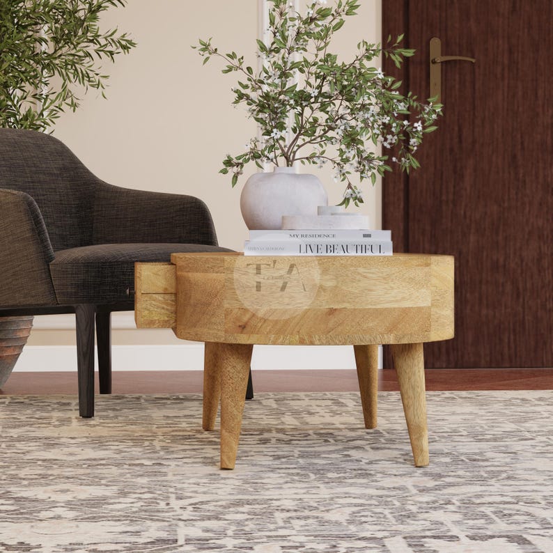 May include: A light wood oval-shaped side table with a drawer and four legs. The table is sitting on a patterned rug in a living room setting. A book titled "Live Beautiful" is on top of the table.