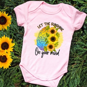 Puede incluir: Un body de beb&eacute; rosa con un gr&aacute;fico de cerebro arco&iacute;ris y el texto "Let the sunshine on your mind".