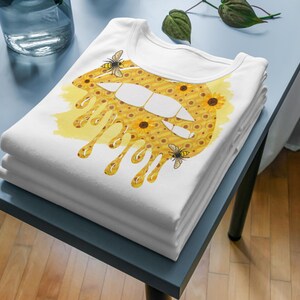 Puede incluir: Una camiseta blanca con un dise&ntilde;o de labios goteantes amarillo y marr&oacute;n. El labio est&aacute; cubierto de peque&ntilde;os girasoles y hay dos abejas en el dise&ntilde;o.