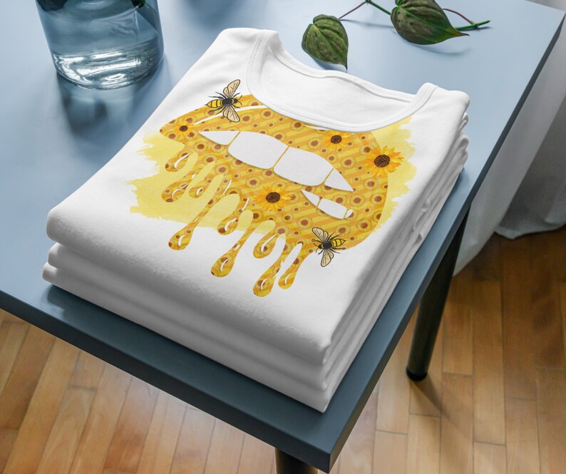 Puede incluir: Una camiseta blanca con un dise&ntilde;o de labios goteantes amarillo y marr&oacute;n. El labio est&aacute; cubierto de peque&ntilde;os girasoles y hay dos abejas en el dise&ntilde;o.