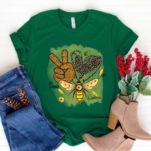 Puede incluir: Camiseta verde con un s&iacute;mbolo de paz, un coraz&oacute;n y un dise&ntilde;o de abeja. El dise&ntilde;o est&aacute; formado por girasoles y las palabras "Peace", "Love" y "Kindness".
