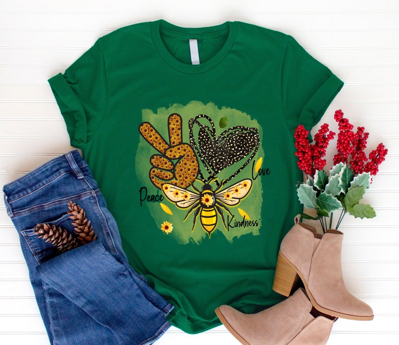Puede incluir: Camiseta verde con un s&iacute;mbolo de paz, un coraz&oacute;n y un dise&ntilde;o de abeja. El dise&ntilde;o est&aacute; formado por girasoles y las palabras "Peace", "Love" y "Kindness".