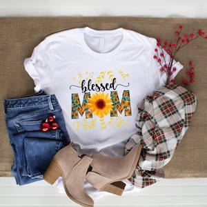 Puede incluir: Camiseta blanca con el texto "blessed mom" en letra negra. Las letras est&aacute;n decoradas con un girasol y un patr&oacute;n de girasol.