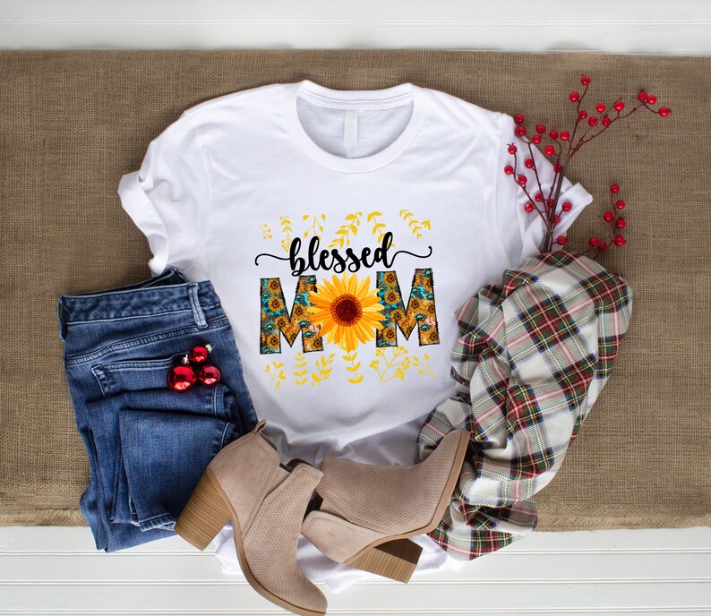 Puede incluir: Camiseta blanca con el texto "blessed mom" en letra negra. Las letras est&aacute;n decoradas con un girasol y un patr&oacute;n de girasol.