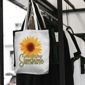 Puede incluir: Bolsa de tela blanca con un girasol amarillo y el texto "You are my sunshine" en letras de purpurina verde.