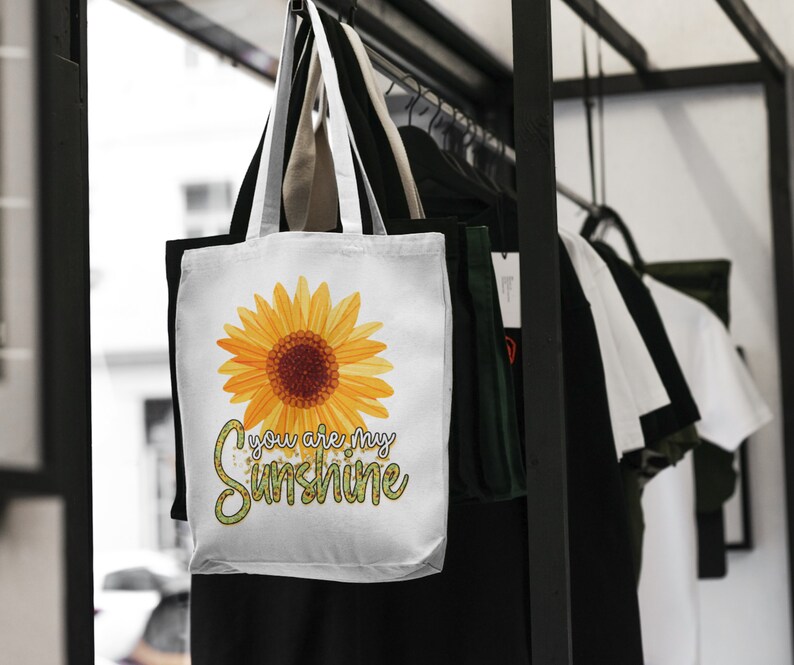 Puede incluir: Bolsa de tela blanca con un girasol amarillo y el texto "You are my sunshine" en letras de purpurina verde.