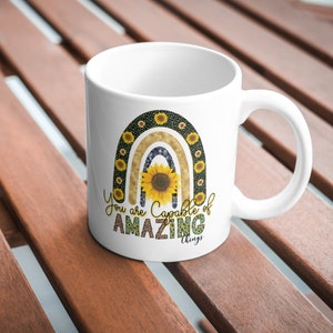 Puede incluir: Taza de cer&aacute;mica blanca con un dise&ntilde;o de arco&iacute;ris con girasoles y el texto "You are capable of amazing things".