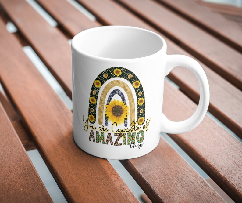 Puede incluir: Taza de cer&aacute;mica blanca con un dise&ntilde;o de arco&iacute;ris con girasoles y el texto "You are capable of amazing things".