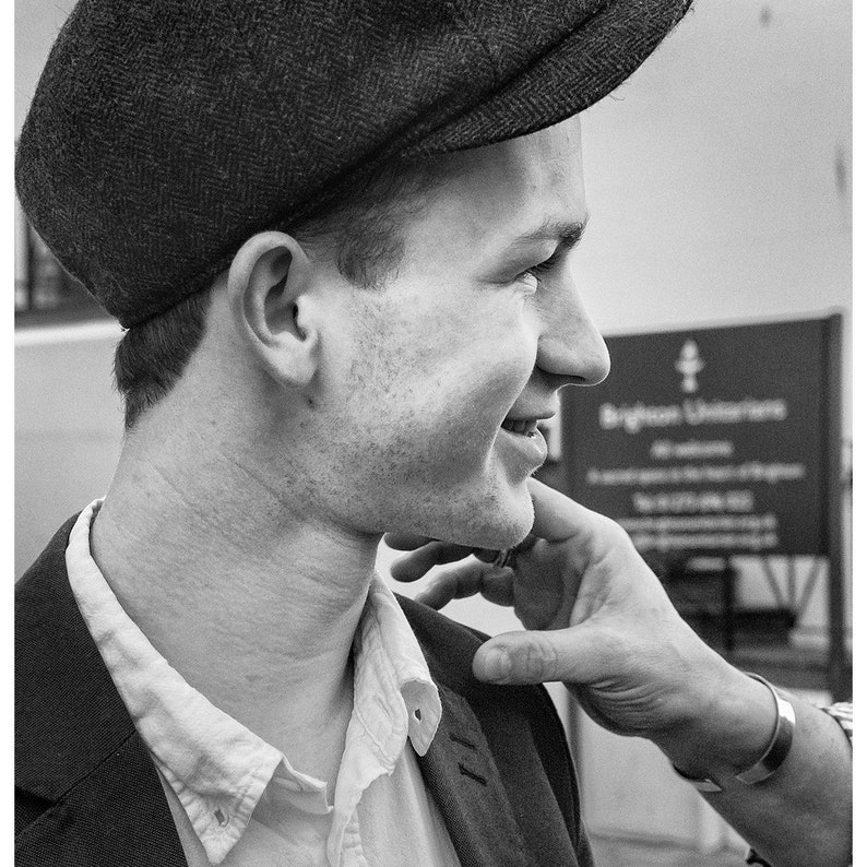 Może przedstawiać: Mężczyzna w tweedowej czapce z daszkiem i białej koszuli z kołnierzykiem pod ciemnym blezerem. Uśmiecha się i patrzy w prawo. Ręka dotyka jego policzka.