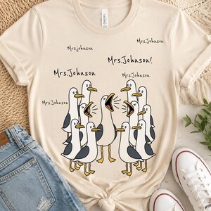 Puede incluir: Una camiseta de manga corta color crema con un gráfico de una bandada de gaviotas blancas y grises con picos y patas amarillas. Las gaviotas aparecen con los picos abiertos y el texto "Mrs. Johnson" se repite varias veces sobre ellas.