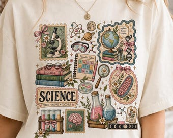 Puede incluir: Una camiseta color hueso con un estampado gráfico colorido de temática científica. El diseño incluye un microscopio, un globo terráqueo, libros, tubos de ensayo, un cerebro, una cadena de ADN y el texto "SCIENCE TEACH INSPIRE DISCOVER".