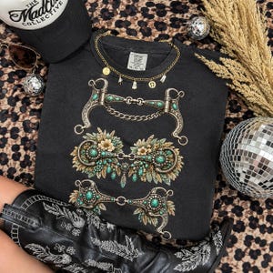 Puede incluir: Una sudadera negra de cuello redondo con un estampado gráfico de tres bocados de caballo ornamentados, decorados con piedras turquesas y detalles florales. Incluye un collar de cadena dorada con pequeños dijes en el cuello.