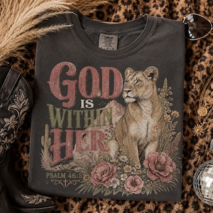 以下が含まれることがあります： チャコールグレーのTシャツで、砂漠の花やサボテンの中に座る雌ライオンのグラフィックプリントが施されています。山を背景に、赤と緑の文字で「God IS WITHIN HER PSALM 46:5」というヴィンテージ風の掠れたフォントのテキストが描かれています。