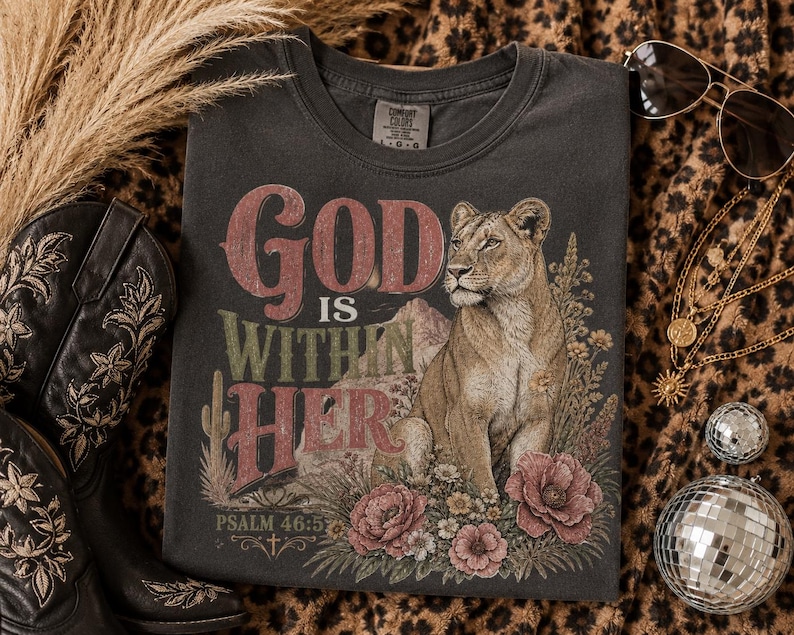 以下が含まれることがあります： チャコールグレーのTシャツで、砂漠の花やサボテンの中に座る雌ライオンのグラフィックプリントが施されています。山を背景に、赤と緑の文字で「God IS WITHIN HER PSALM 46:5」というヴィンテージ風の掠れたフォントのテキストが描かれています。