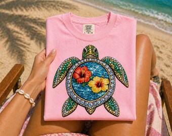 May include: A light pink t-shirt featuring a detailed sea turtle design. The turtle has a blue and turquoise shell with a red and yellow flower design. The shirt has the words "Comfort Colors" on the tag.
