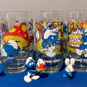 Puede incluir: Juego de seis vasos transparentes con personajes de los Pitufos, incluyendo Pitufo Torpe, Perezoso, Manitas, Panadero y Douchy. Cada vaso tiene una ilustración diferente de los Pitufos. Pequeñas figuras de los Pitufos están dispuestas frente a los vasos.