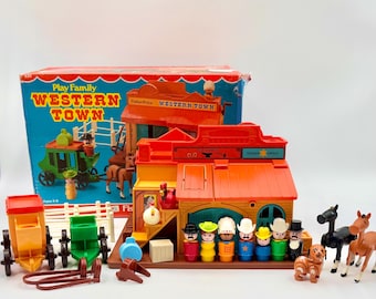 May include: A vintage Fisher-Price Play Family Western Town playset. The set includes a Western-style town building, a stagecoach, horses, and several cowboy and townsperson figures. The box is visible in the background, with the words "Western Town" prominently displayed.
