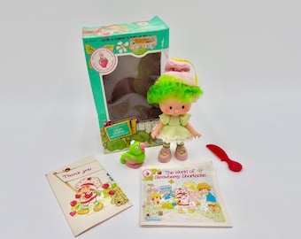 Puede incluir: Una muñeca Lime Chiffon Strawberry Shortcake con cabello verde brillante, sombrero rosa y vestido verde claro. Incluye una figura de pájaro verde, un peine de plástico rojo, una caja de cartón con texto, una tarjeta de agradecimiento y un folleto titulado The World of Strawberry Shortcake.