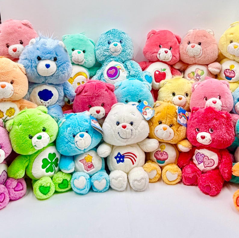 May include: A collection of colorful plush teddy bears in various shades including blue, pink, green, yellow, and white. Each bear features a unique embroidered symbol on its belly, white heart-shaped paw pads, and a friendly facial expression.