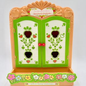 May include: A peach-colored plastic toy cabinet with two white doors featuring green strawberry-shaped cutouts and red knobs. The top arch reads The Wonderful World of Strawberry Shortcake, and the base features a floral border with the text Life is Berry Delicious.