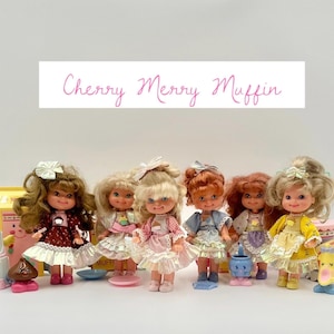 Może przedstawiać: Kolekcja lalek Cherry Merry Muffin w stylu vintage, każda z unikalnymi strojami i fryzurami. Lalki otoczone są miniaturowymi akcesoriami, w tym ciastami i ciastem. Tekst "Cherry Merry Muffin" jest wyświetlany nad lalkami.