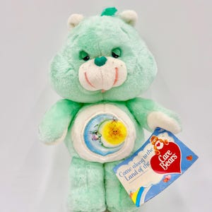 May include: A mint green Care Bear plush toy with a smiling face, a heart-shaped nose, and a belly badge featuring a moon and sun. The plush toy has a tag that reads "Come along to the Land of the Care Bears."