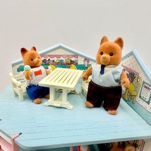 May include: A miniature cafe playset featuring two brown bear figures. One bear holds an ice cream cone, while the other wears a shirt and tie. The set includes a table, chairs, and a decorative backdrop with floral and menu details.