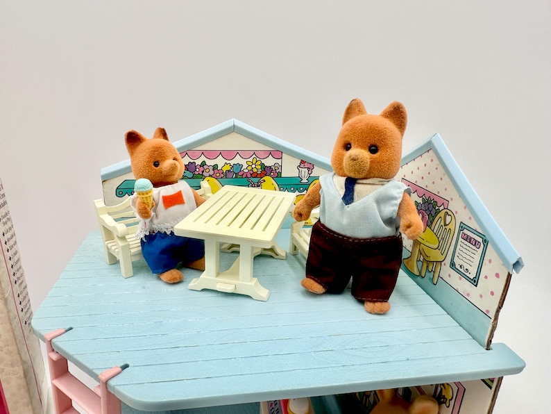 May include: A miniature cafe playset featuring two brown bear figures. One bear holds an ice cream cone, while the other wears a shirt and tie. The set includes a table, chairs, and a decorative backdrop with floral and menu details.