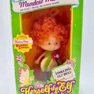 May include: Vintage "Herself the Elf & Friends" doll in original packaging. The doll, "Meadow Morn," has red curly hair and wears a green and yellow dress. The box is green with text including "Tinkling Elf Bell" and "Bluebird Charm."