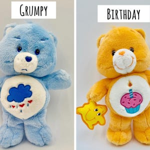 May include: Two plush bears. The left bear is light blue with a white belly patch featuring a blue rain cloud and red hearts, labeled GRUMPY. The right bear is orange with a white belly patch featuring a cupcake with a candle, labeled BIRTHDAY.