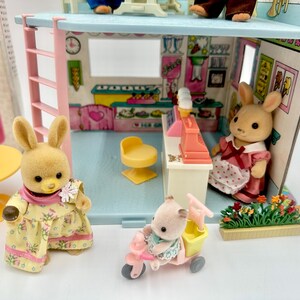 May include: A miniature toy shop playset with bunny figurines. The shop features a counter, a yellow chair, and a pink cash register. One bunny wears a yellow dress, another a pink dress, and a third rides a pink tricycle with a yellow basket.