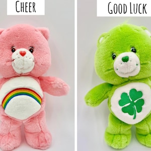May include: Two plush bears. The left bear is pink with a white belly patch featuring a rainbow and the text Cheer above it. The right bear is lime green with a white belly patch featuring a four-leaf clover and the text Good Luck above it.