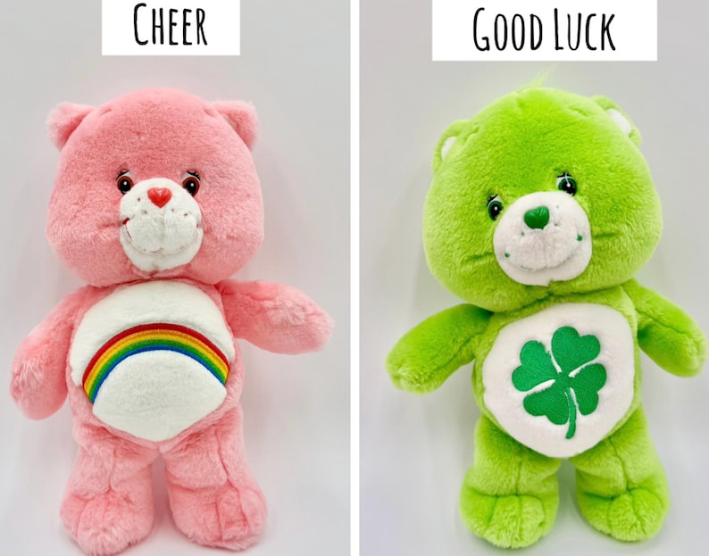 May include: Two plush bears. The left bear is pink with a white belly patch featuring a rainbow and the text Cheer above it. The right bear is lime green with a white belly patch featuring a four-leaf clover and the text Good Luck above it.